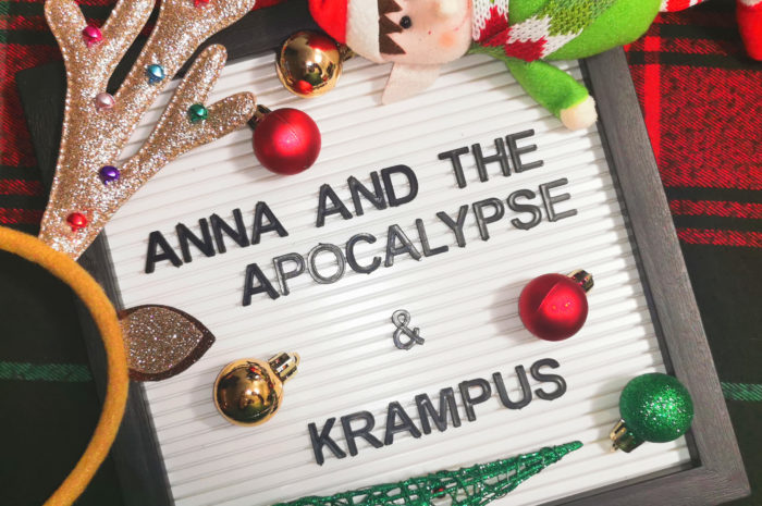 A sign that says Anna and the Apocalypse and Krampus surrounded by a stuffed elf, sparkly antlers, and a wire Christmas tree with a plaid background